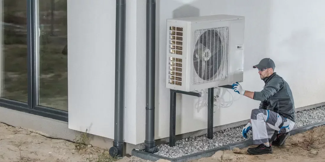 A man in grey kneels down next to an outdoor heat pump unit on the side of a white building