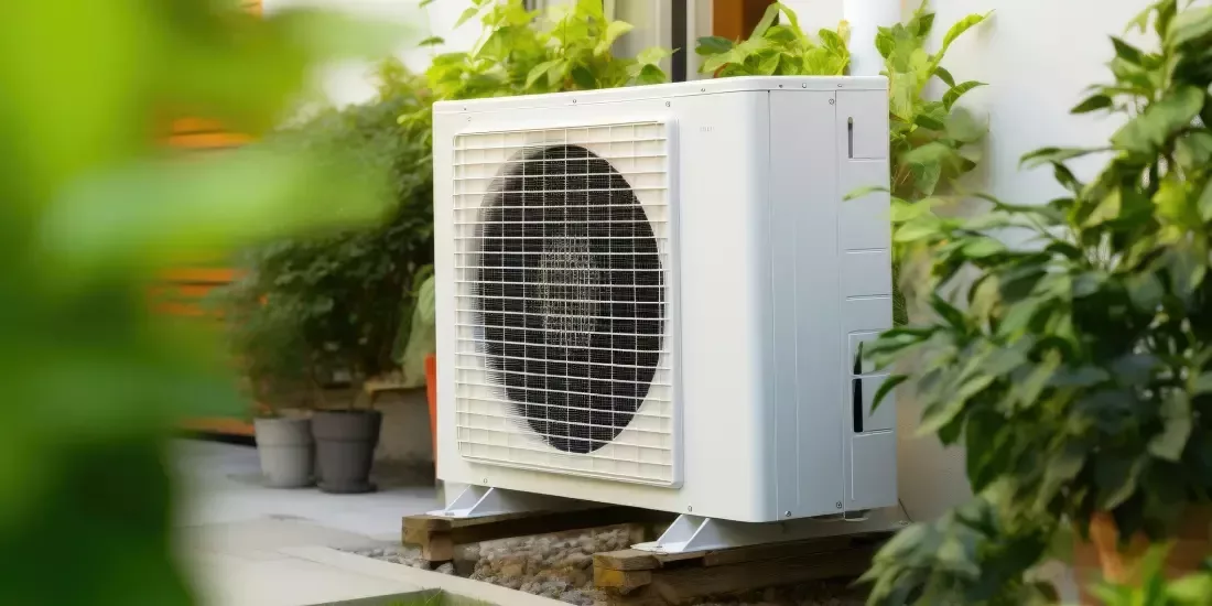 A white air source heat pump unit outside surrounded by green leaves