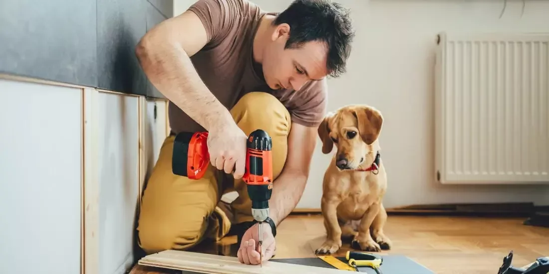 A man drills a screw into a piece of wood while a dog watches him