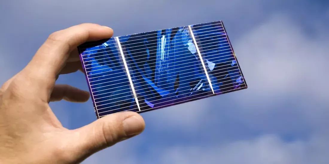 A hand holding up a blue solar cell against a cloudy sky