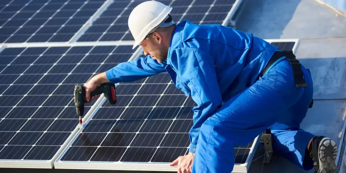 A man in a blue uniform uses a drill to secure a solar panel to a residential roof