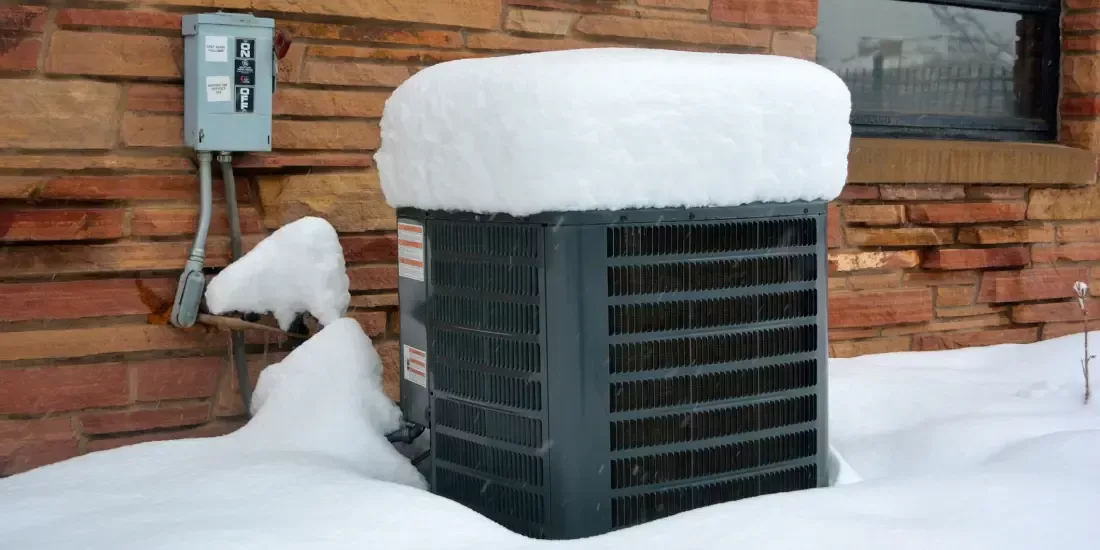 A heat pump covered in snow outside a brown bricked building